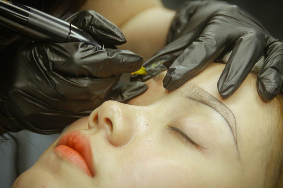 A detailed view of eyebrow microblading procedure with gloved hands and closed eyes. - Photo by Thanh Cong Le on Pexels